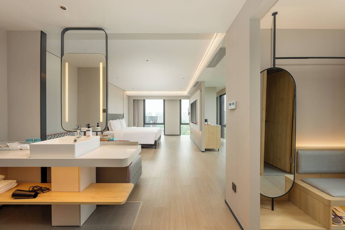 Una habitación de hotel moderna con una cama grande, una mesita de noche, un escritorio con una computadora, un espejo amplio y vistas a la ciudad a través de las ventanas.