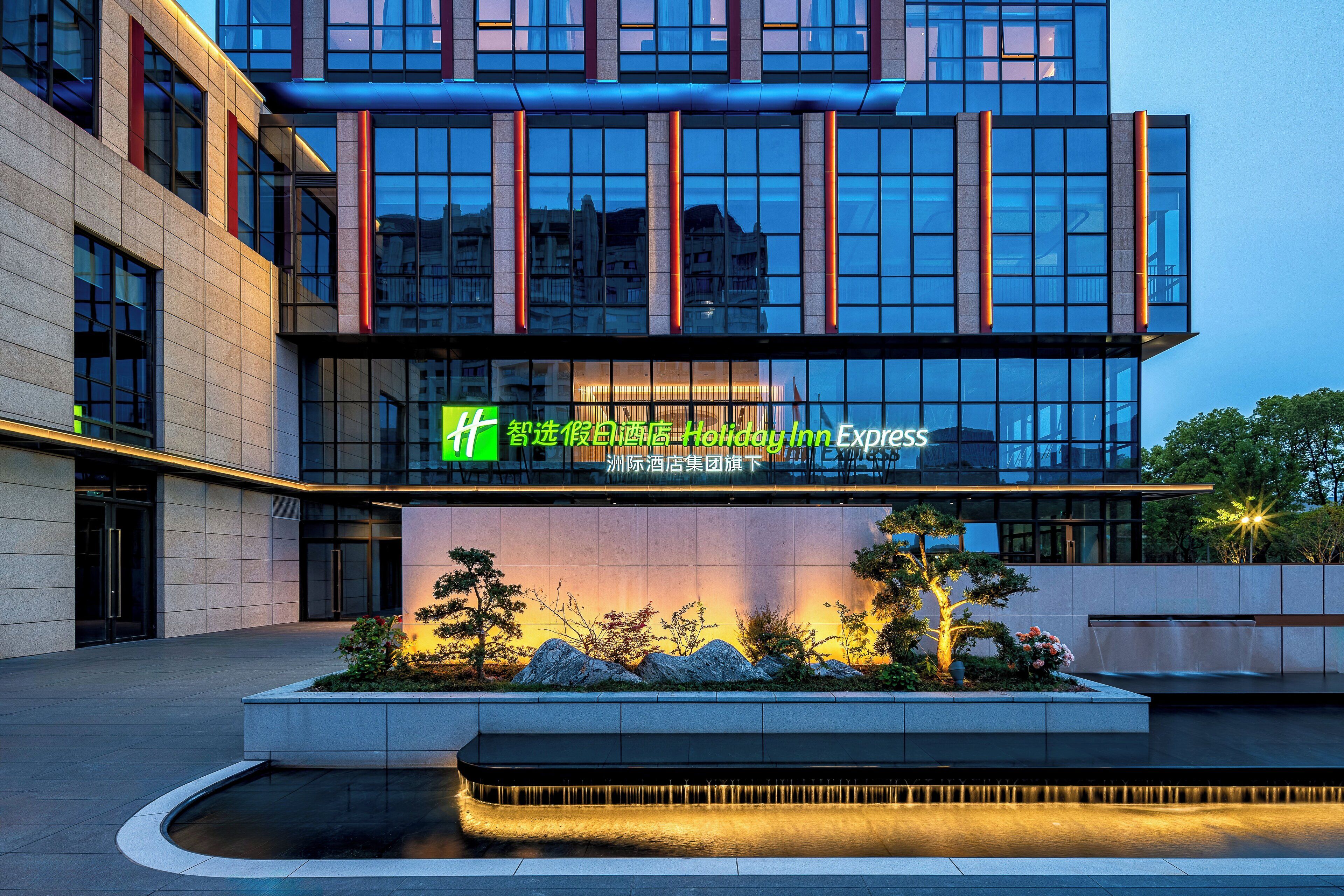 A modern hotel building with a Holiday Inn Express sign, featuring a landscaped garden with rocks and trees, and a water feature in front.