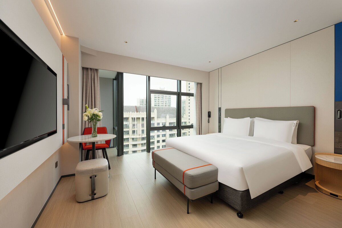 Habitación de hotel moderna con una cama grande, un escritorio con una silla, una mesita con un florero y un ventanal con vistas a la ciudad.
