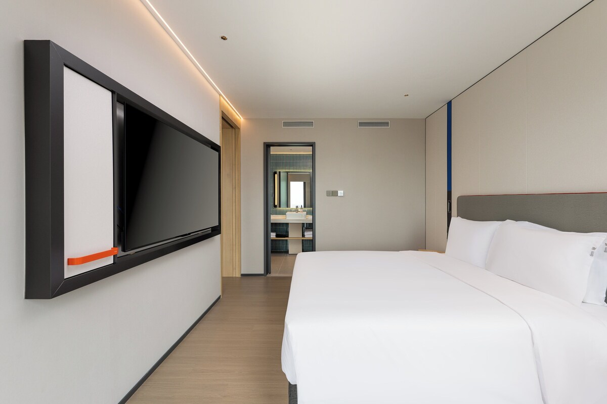 Una habitación de hotel moderna con una cama grande, un televisor de pantalla plana montado en la pared y un baño visible a través de una puerta abierta.
