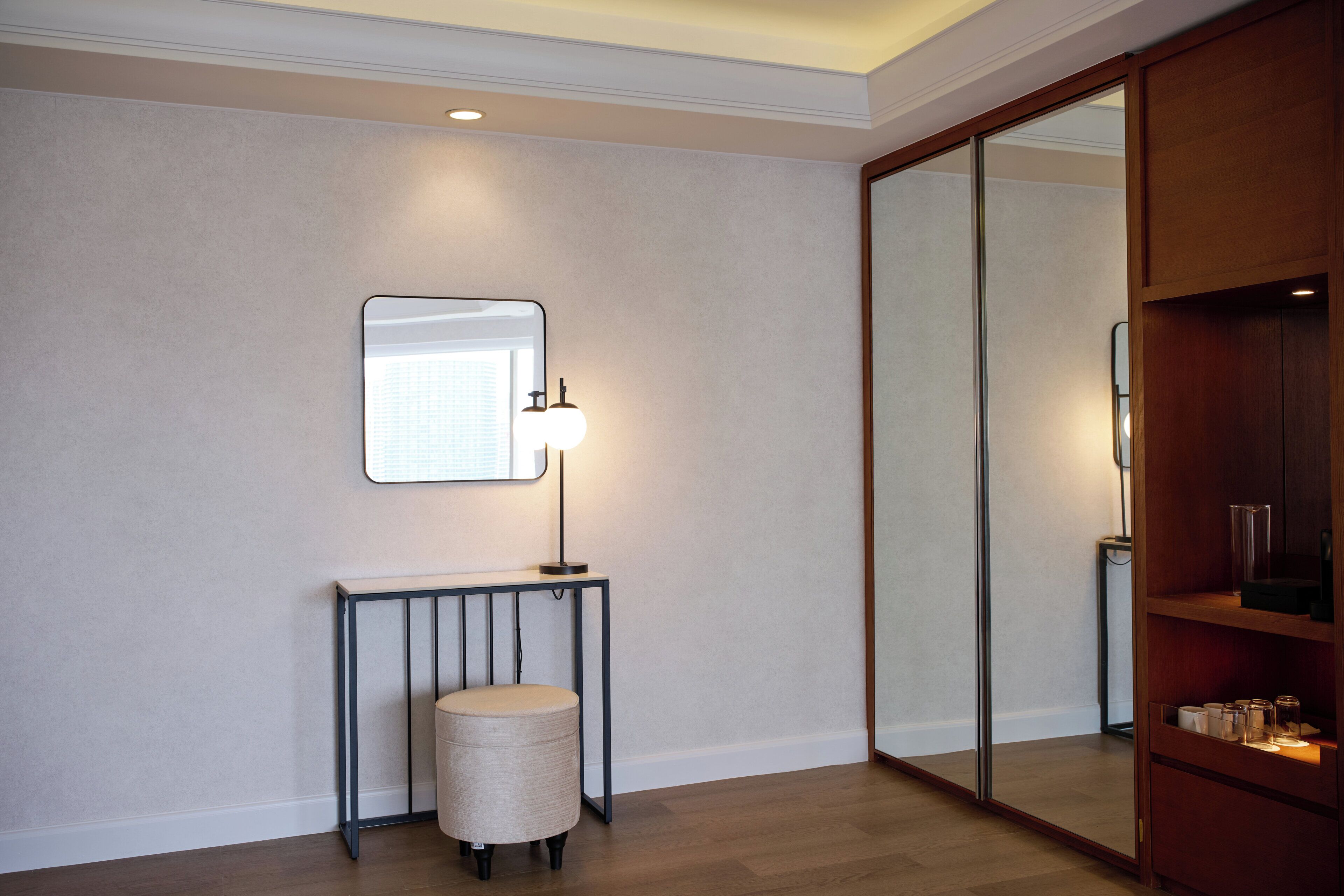 A room with a mirror, a table with a lamp, a round ottoman, and a wooden wardrobe.