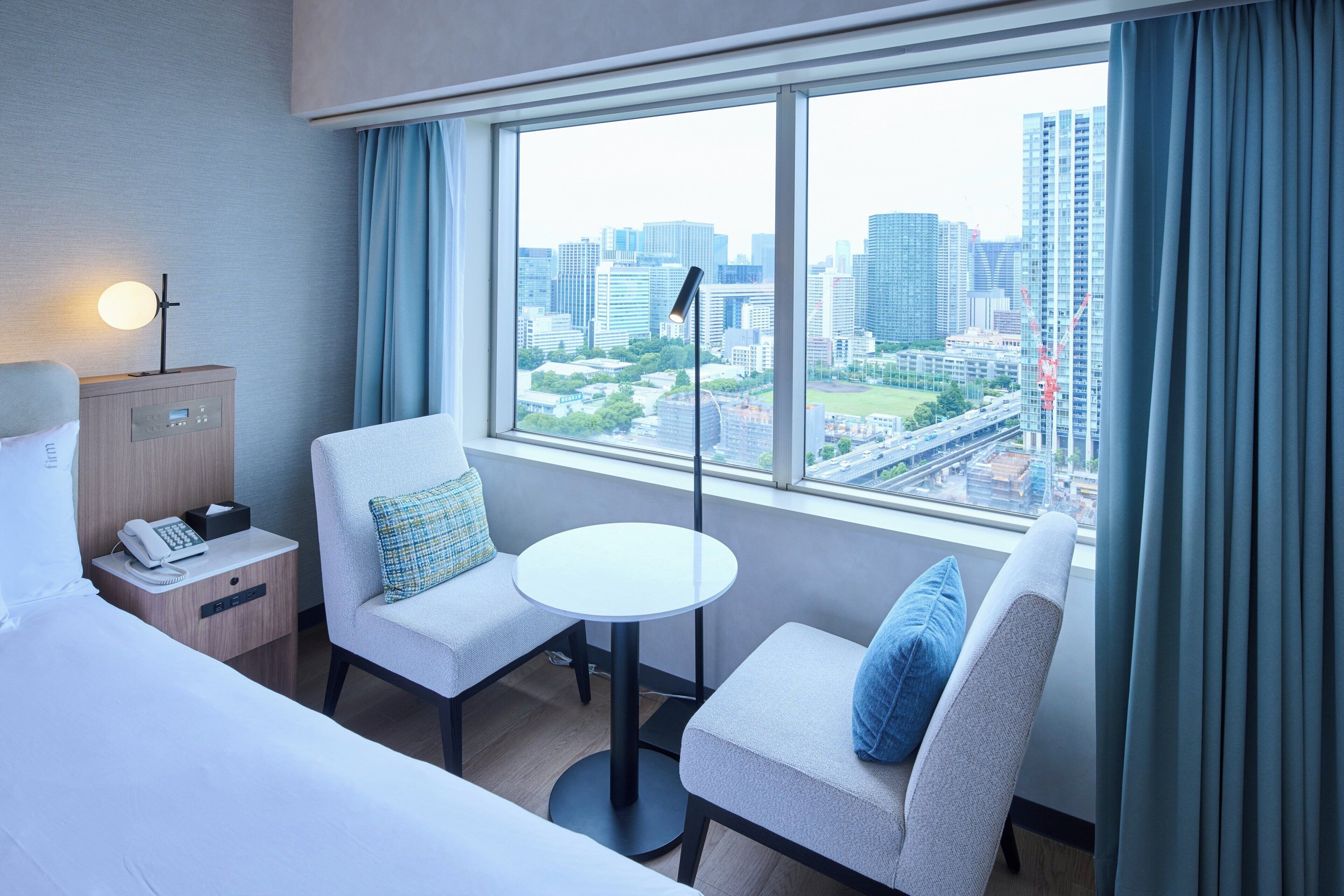 A hotel room with a bed, two armchairs, a small table, a bedside table with a lamp, a phone, and a view of a cityscape through large windows.
