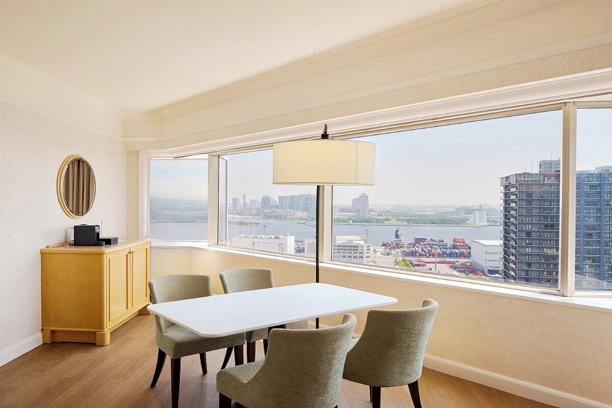 Une chambre d’hôtel moderne dotée d’une grande fenêtre donnant sur la ville, d’une table blanche avec quatre chaises et d’un meuble en bois avec un miroir.