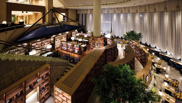 Lobby d'albergo su più livelli con un atrio centrale, scaffali di libri, zone relax e una scala.