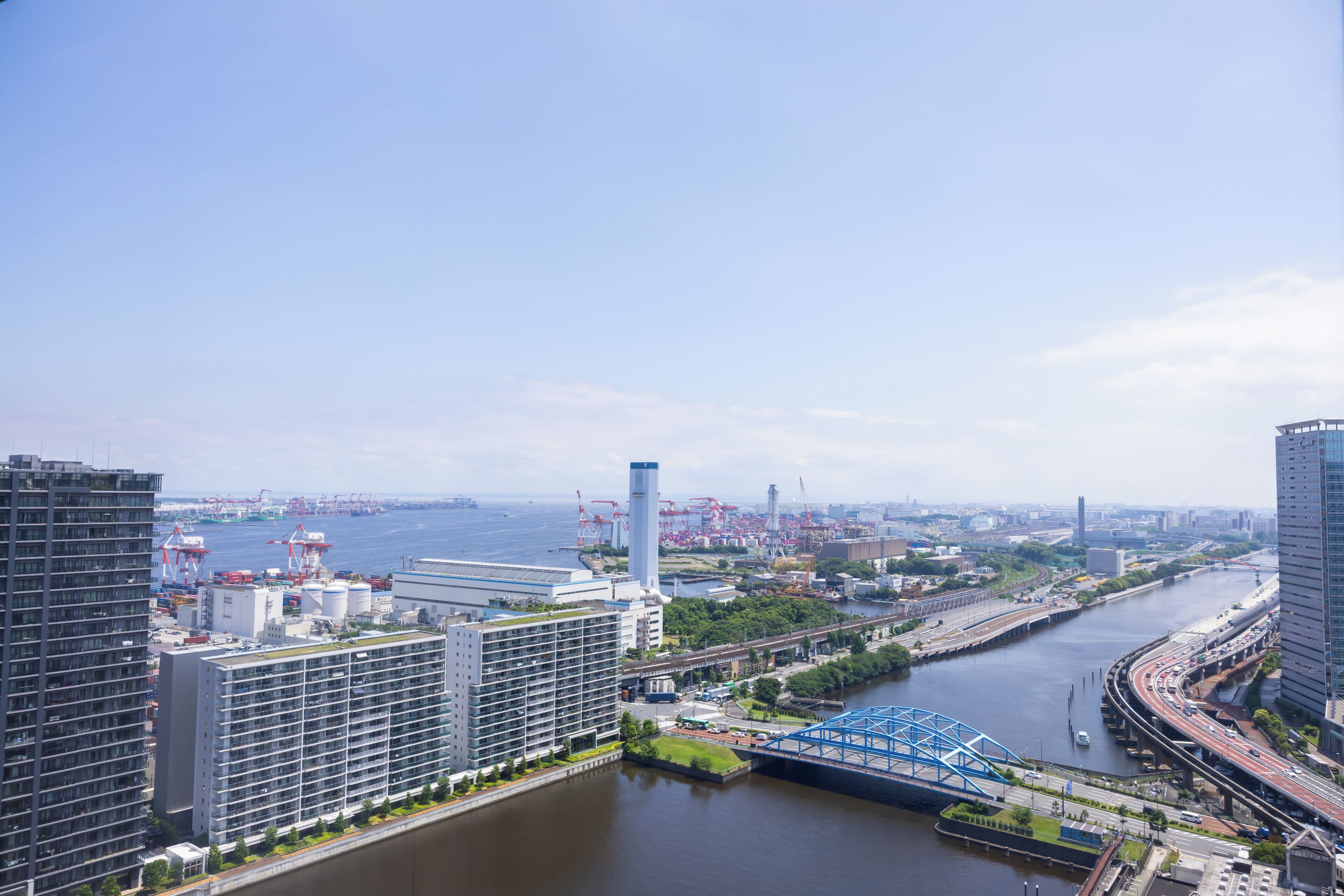 A high-rise building with a modern design, a river with a bridge, and a distant view of a port with cranes.