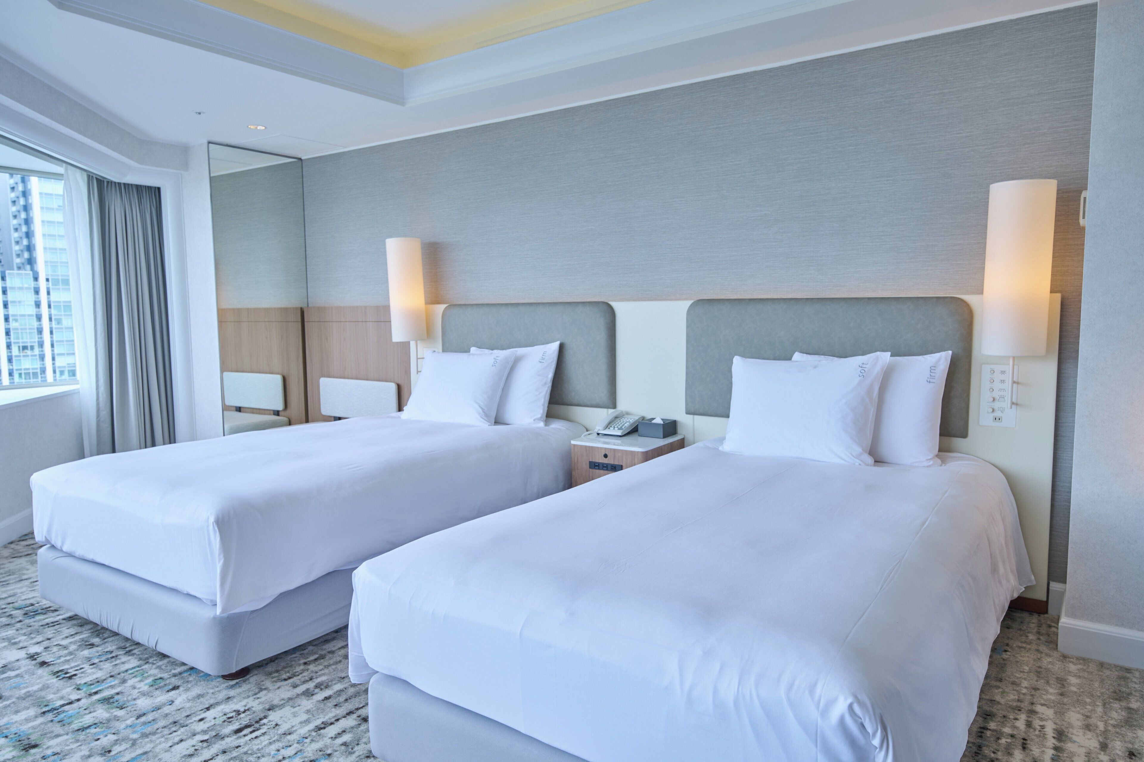 A hotel room with two beds, a large window with curtains, a bedside table with a lamp, and a wall-mounted phone.