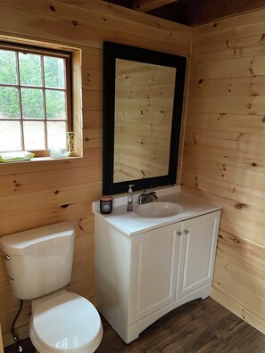 A small, wooden cabin bathroom with a white toilet, a sink with a mirror above it, and a window with a view of trees. - Rustic and Sustainable mini Cabin (i.e. no electricity) (Belgrade)