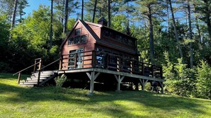 A cabin with a wooden deck and stairs leading up to it, surrounded by tall trees.