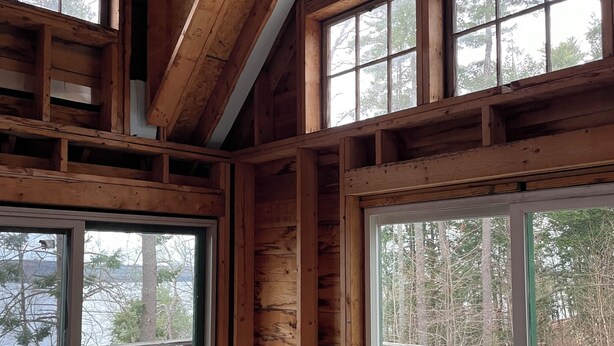 A wooden cabin with a couch, pillows, and a view of a forest and lake.