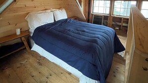 A wooden loft bedroom with a sloped ceiling, a bed with a blue and white duvet, and a window overlooking a forest.