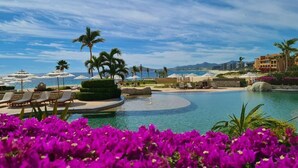Zona de piscina en un resort con sillas de descanso, palmeras y vista despejada a la playa y al océano.