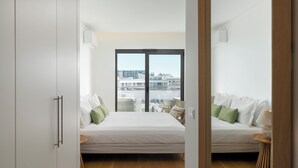 A modern bedroom with a large bed, a small side table, and a view of the city through a large window. - With shared swimming pool and gym - Acqua Residence IV (Funchal)