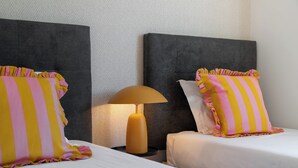 Une chambre d’hôtel avec deux lits, chacun agrémenté d’un oreiller à rayures jaunes et roses. Une table de chevet avec une lampe se trouve entre les deux lits.