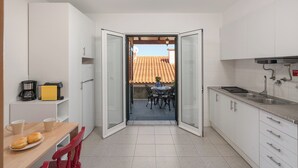 Une cuisine compacte avec des meubles blancs, un coin repas comprenant une table et des chaises, et une vue sur un toit recouvert de tuiles, visible à travers une porte ouverte.