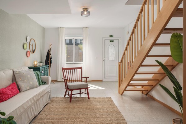A modern living room with a wooden staircase, a sofa with patterned cushions, a wooden chair, and a large plant.
