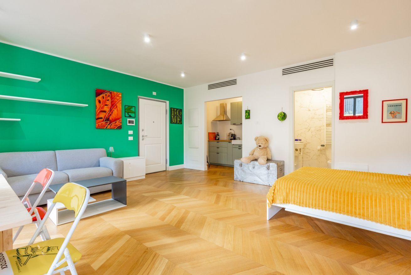 A room with a green wall, a sofa, a bed with a yellow blanket, a teddy bear, and a kitchen area in the background.