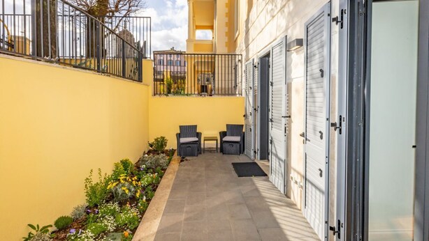 Un étroit couloir extérieur avec un mur jaune, deux chaises en osier, une petite table et un portail en métal.