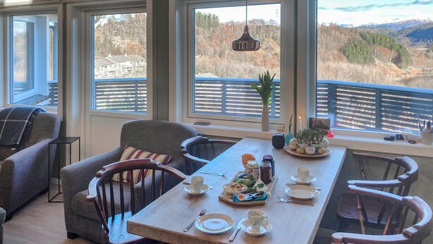 A dining area with a wooden table set for four, surrounded by chairs. Large windows offer a view of a mountainous landscape. - Lovely home in Flaktveit with WiFi (Flaktveit)