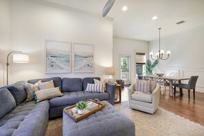 Uma sala de estar com um sofá seccional cinza, uma mesa redonda pequena e um espaço de jantar com mesa e cadeiras de madeira.