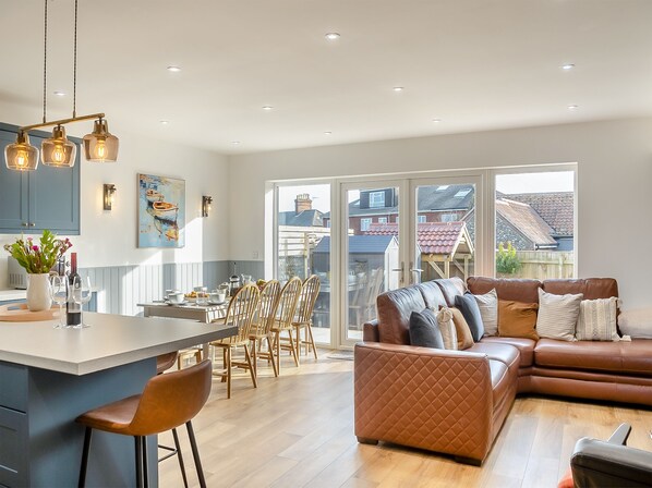 A modern kitchen with a central island, dining area with chairs, a brown leather sofa, and a large window with a view of houses.