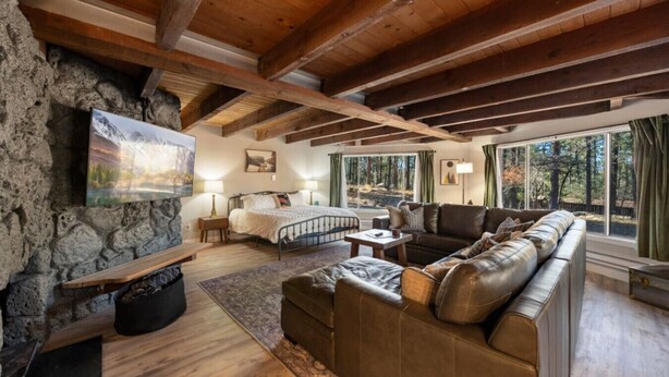 A cozy cabin interior with a stone wall, wooden beams, a large leather sofa, a bed, and a fireplace. - Morning Bliss - Multi Family & Couples (Prescott)