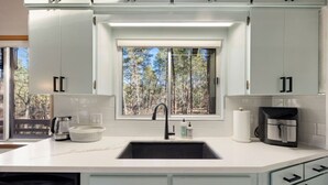 A modern kitchen with white cabinets, a built-in sink, and a window overlooking a forest. - Morning Bliss - Multi Family & Couples (Prescott)