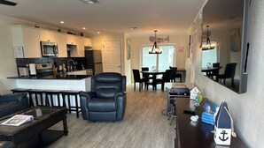 A modern living room with a black leather armchair, a glass coffee table, and a wooden dining table with chairs. A kitchen with white cabinets and a microwave is visible in the background. - Sunset By The Sea 3052 - FMB, Family Friendly (Fort Myers Beach)