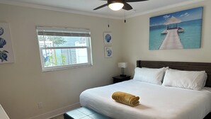 A neatly arranged bedroom with a bed, a bench, a nightstand, a ceiling fan, a window with blinds, and a painting of a pier. - Sunset By The Sea 3052 - FMB, Family Friendly (Fort Myers Beach)