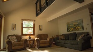 A spacious living room with a high ceiling, a skylight, and a large window. There are two sofas, a coffee table, and a chair.
