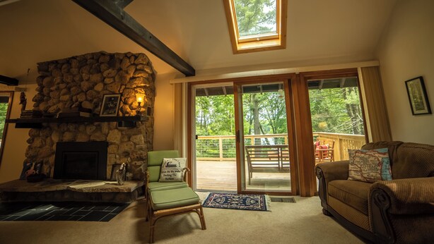 A cozy living room with a stone fireplace, a comfortable green chair, a brown sofa, and a large window overlooking a deck.