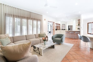 A modern living room with a beige sofa, a marble coffee table, and a grey armchair. A kitchen with white cabinets and a bar area is visible in the background.