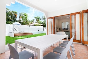 A modern outdoor patio with a white table and chairs, a barbecue area, and a view of a lush garden with palm trees.
