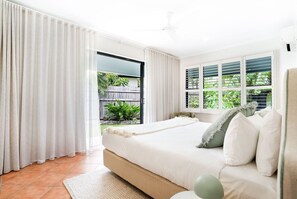 Ein Schlafzimmer mit einem großen Bett, einem Fenster mit Jalousien, einem Deckenventilator und Blick auf Grünfläche.