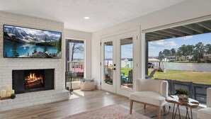 A modern living room with a fireplace, a large TV mounted on a brick wall, and a view of a lake and mountains through large glass doors. - 4\2.5+ LAKE HOUSE — Fish & Swim @DOCK; LakeMat; Kayak; PaddleBoard; King & Queen (Montgomery)