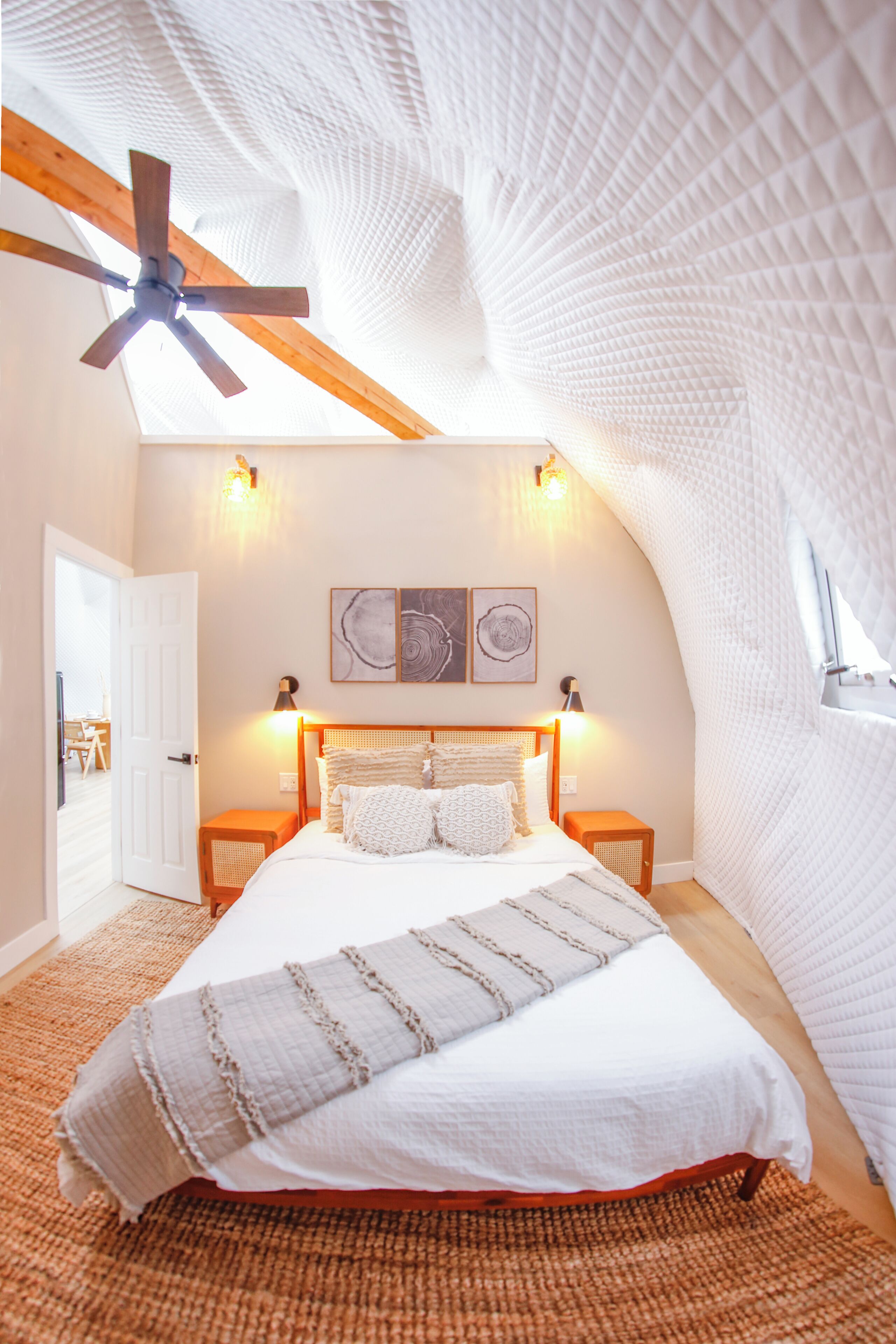 Quarto com teto inclinado, ventoinha de madeira, cama com roupa de cama branca, duas luminárias de cabeceira e uma obra de arte na parede acima da cama.