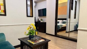 Una sala de estar con un sofá color turquesa, una mesa de centro con un jarrón de girasoles, y un área de cocina visible a través de una puerta abierta.