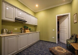 A compact kitchen with white cabinets, a built-in stove, and a patterned floor. A door leads to a living area with a patterned rug and a bench. - Seven Gate Suit Hotel (Baku)