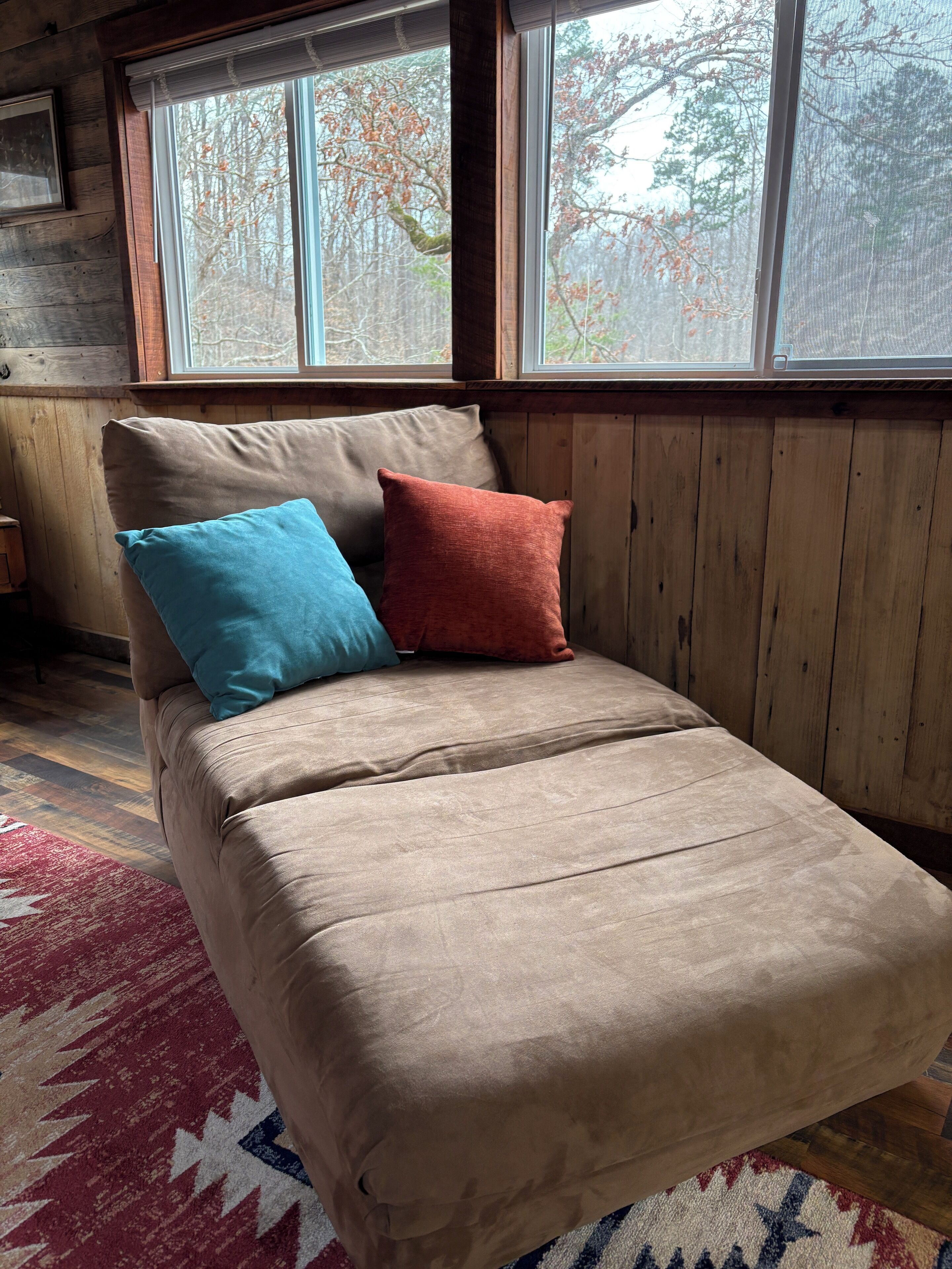 A bed with a beige cover and two pillows, one blue and one red, in a room with wooden walls and large windows.