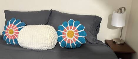 A bed with dark gray sheets, two crocheted pillows with floral patterns, and a bedside table with a lamp.