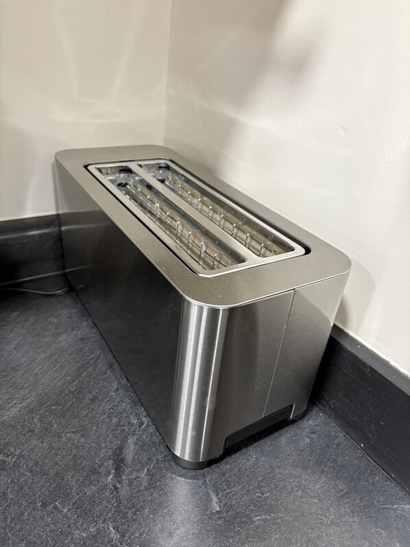 Ein Edelstahl-Toaster mit schwarzem Sockel steht auf einer dunklen Arbeitsplatte vor einer weißen Wand.