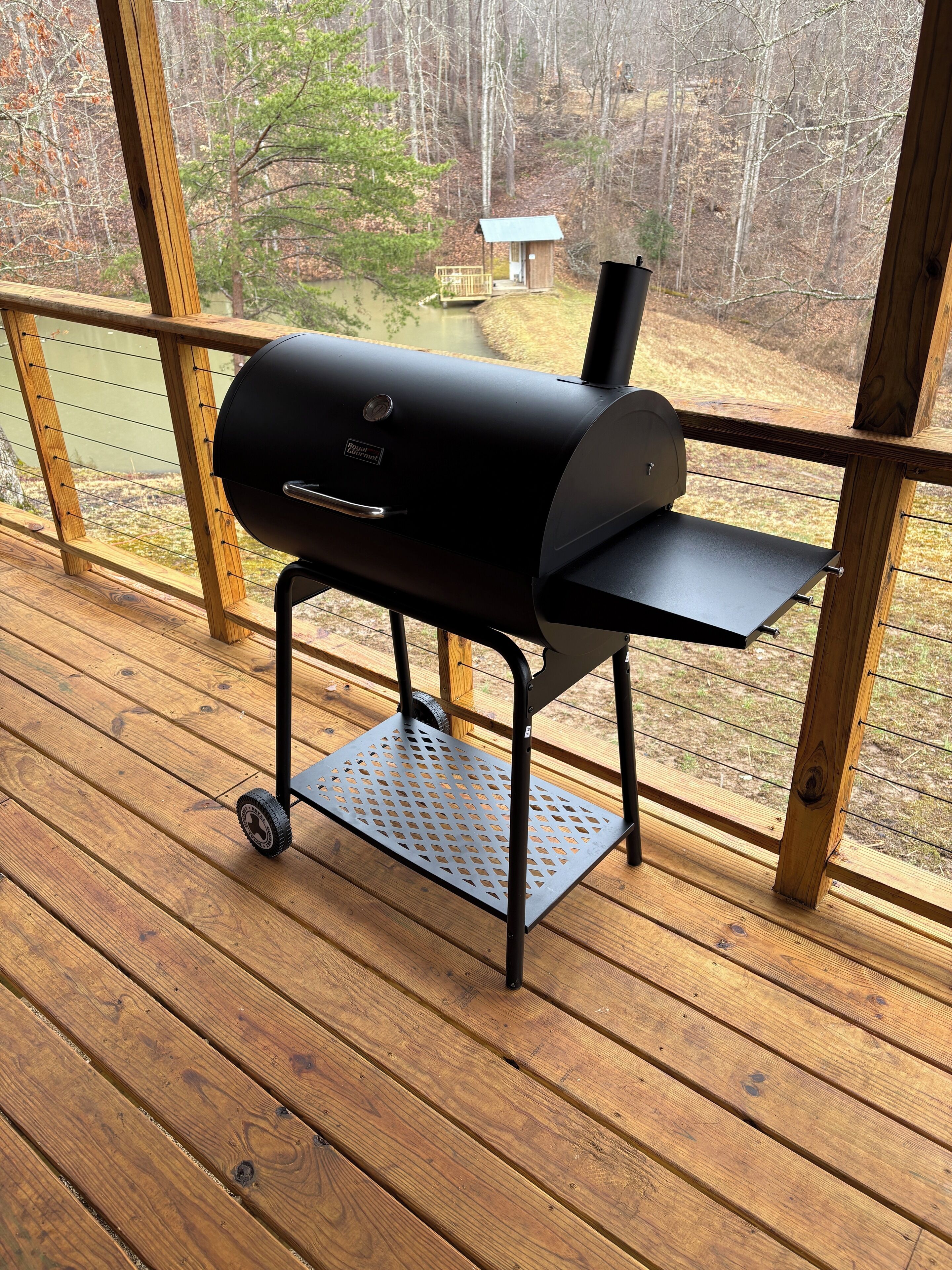 A portable grill on a wooden deck with a view of a forest and a small cabin.