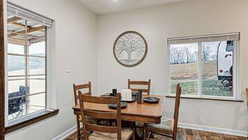 A dining area with a wooden table and chairs, a decorative tree wall art, and a window with a view of a vehicle and landscape.