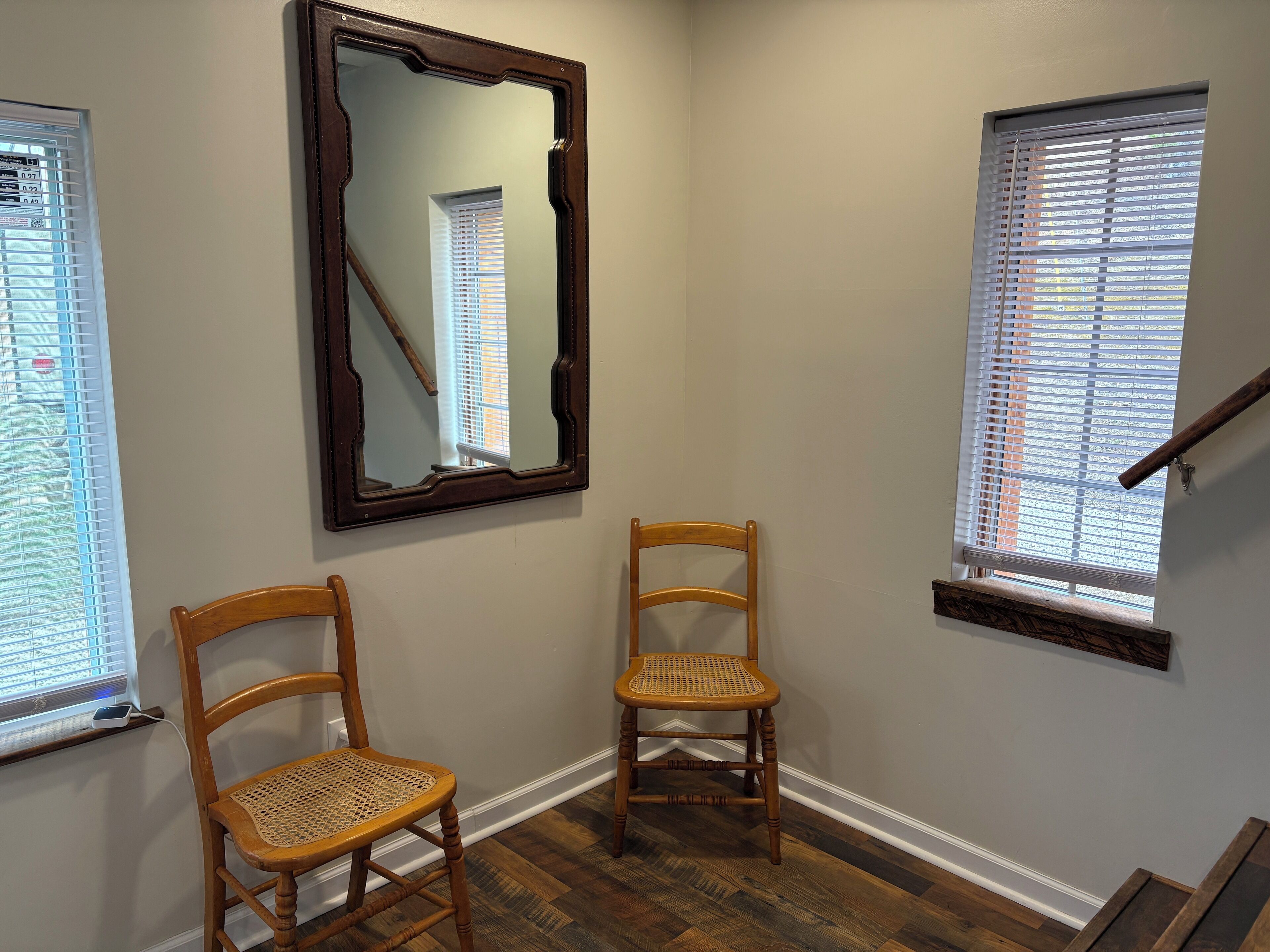 A room with two wooden chairs, a large mirror, and two windows with blinds.