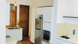 Een moderne keuken met witte kastjes, een roestvrijstalen koelkast en een inbouwoven. Er is een houten deur en een wit aanrecht met een spoelbak.