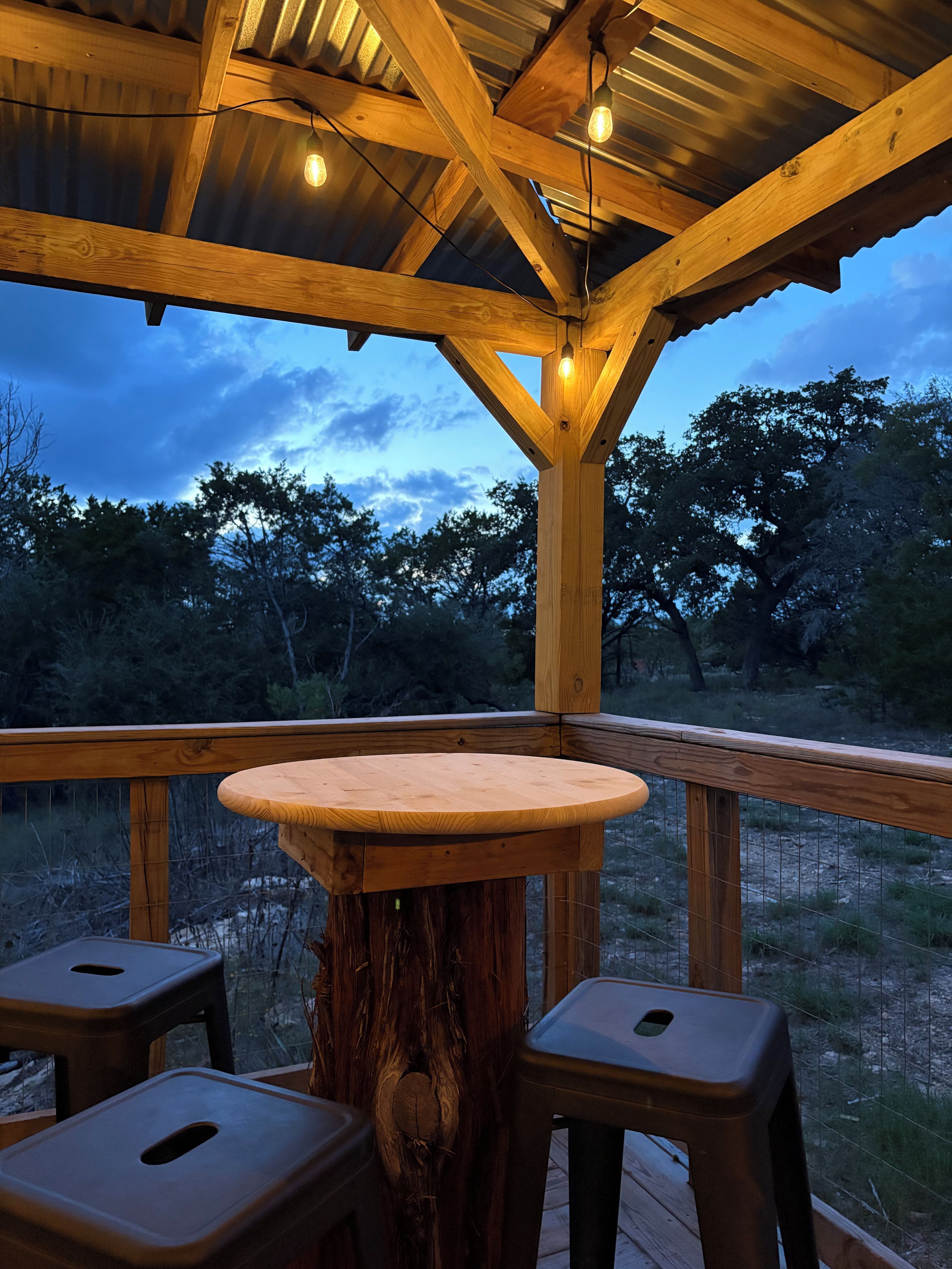 An outdoor wooden deck with a round table and two stools, surrounded by a metal railing.