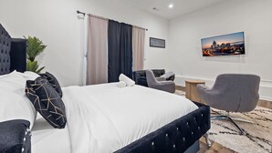 A modern hotel room with a bed, a chair, a small table, and a cityscape painting on the wall. - Atlanta Game House with Hot Tub (Riverdale)
