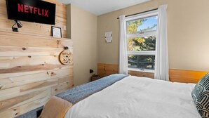 A bedroom with a bed, a wooden wall, a window with a view, and a TV mounted on the wall. - Cabin on Mtn, Seasonal Pool Access w Panoramic Views! Hike, Golf, Swim, Unwind! (Lanesville)