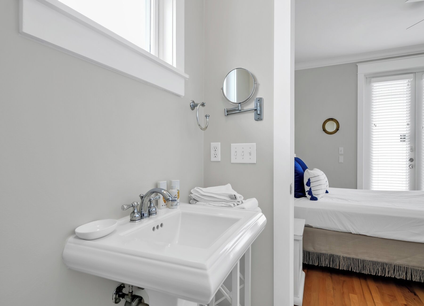 A bathroom with a sink, mirror, and towel rack. There is a window with blinds and a bed in the adjacent room.