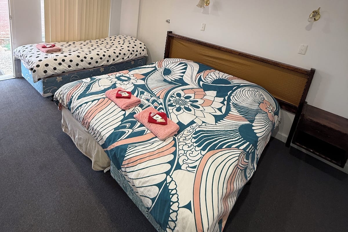 A hotel room with two beds, one with a polka dot and one with a floral pattern. There are ceiling lights, a wall-mounted light fixture, and a door with a curtain.