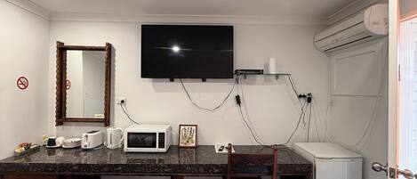 A compact room with a kitchenette, a flat-screen TV mounted on the wall, a wooden chair, a small refrigerator, and a trash bin.